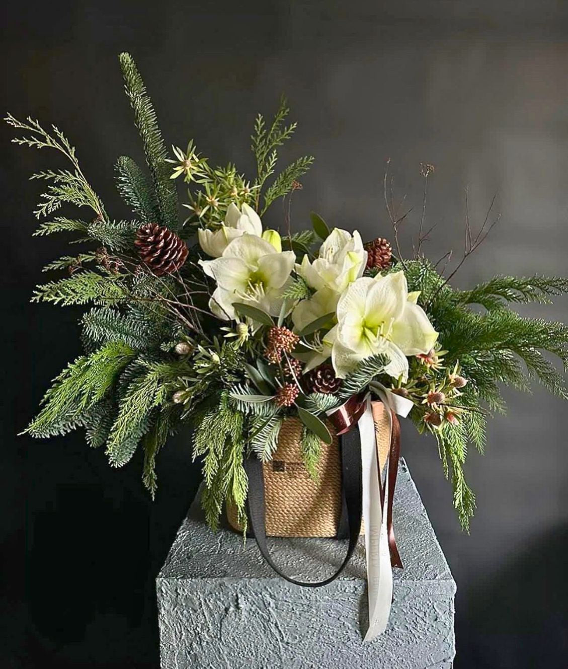 White amaryllis and mixed evergreen in a woven basket, a holiday arrangement by Alaric Flower Design in New York City