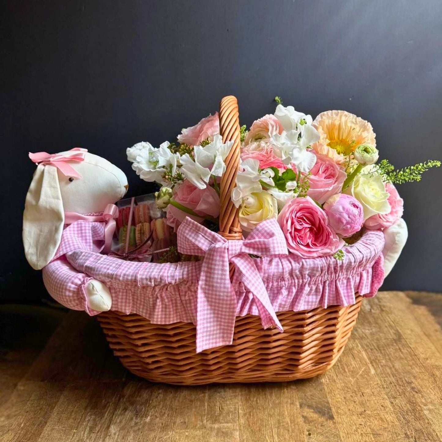 Wicker basket with flowers, a plush bunny, and candy on a wooden surface.