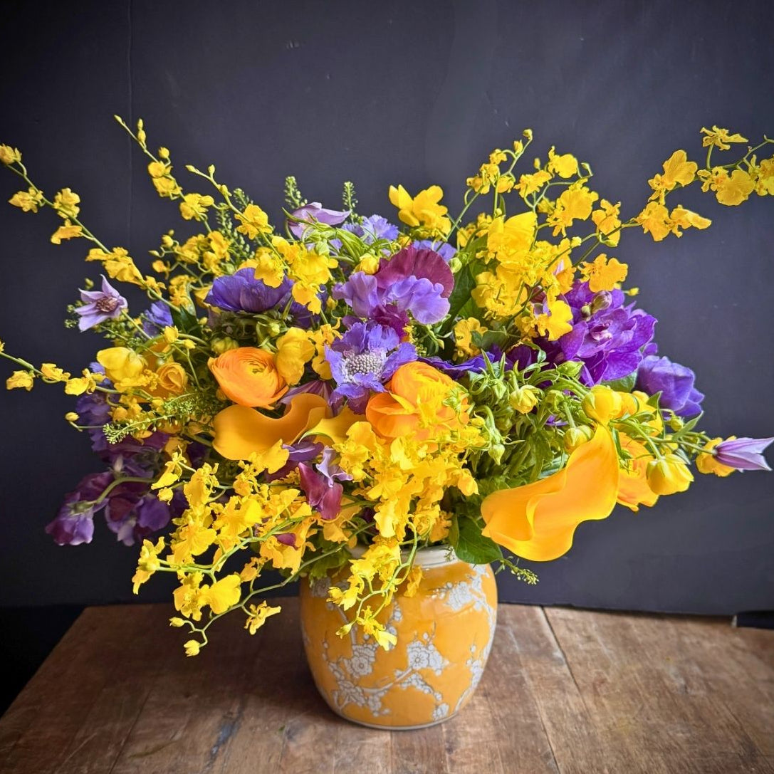A vibrant floral arrangement featuring yellow, purple, and orange flowers in a patterned yellow-orange ceramic vase, set on a rustic wooden table against a dark gray background.